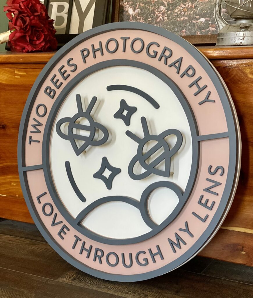 Round logo sign that says two bees photography. Pink, white, and grey. Has two floating bees.