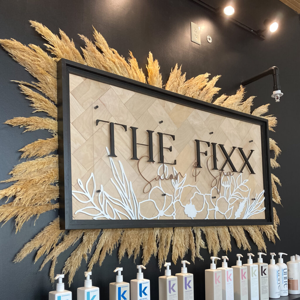 Large framed sign on a black wall with pampas grass behind it. The sign says “the fixx salon & spa” Shampoo and conditioner bottles line the shelf beneath it